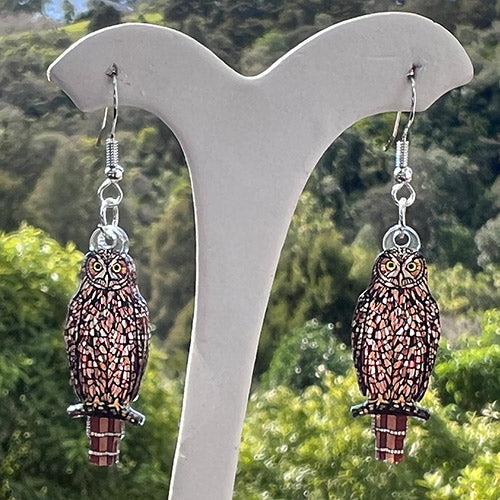 Morepork earring on a white stand with a blurred green background